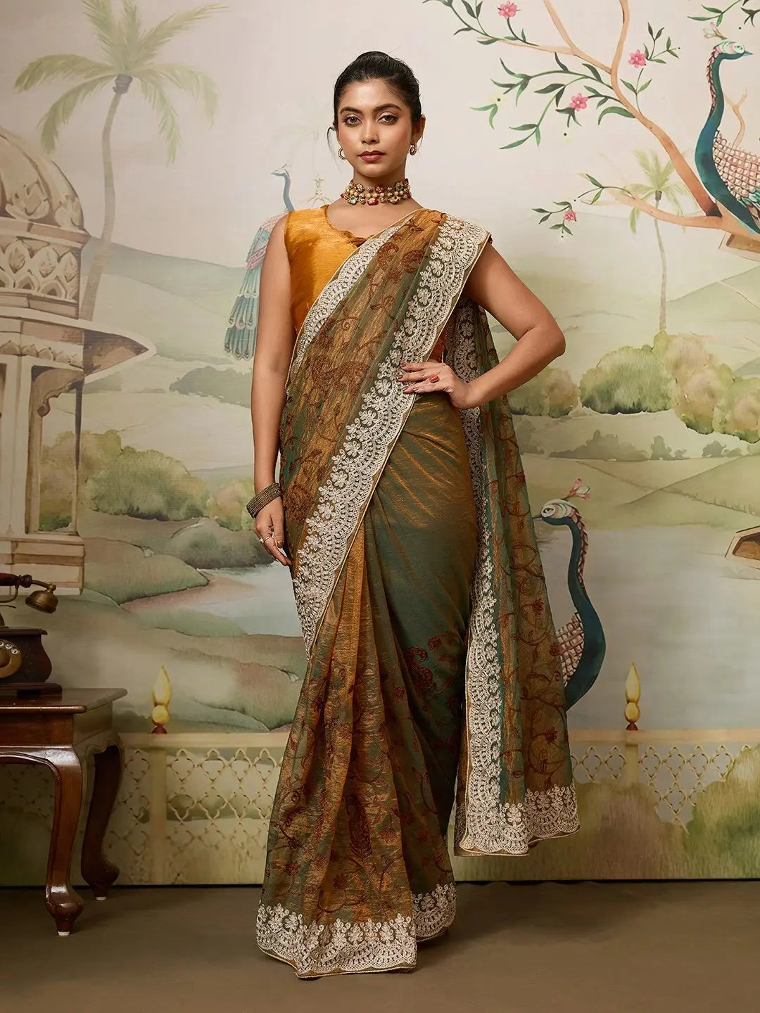 Woman in a traditional saree standing in front of a decorative wall with peacock designs.